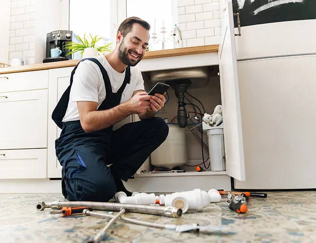 Plombier souriant utilisant son téléphone en réparant l'évier de la cuisine à Longnes dans les Yvelines 78