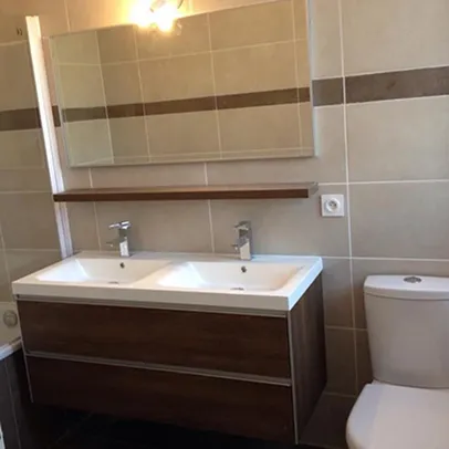 Salle de bain moderne avec double vasque, miroir, WC et carrelage beige.
